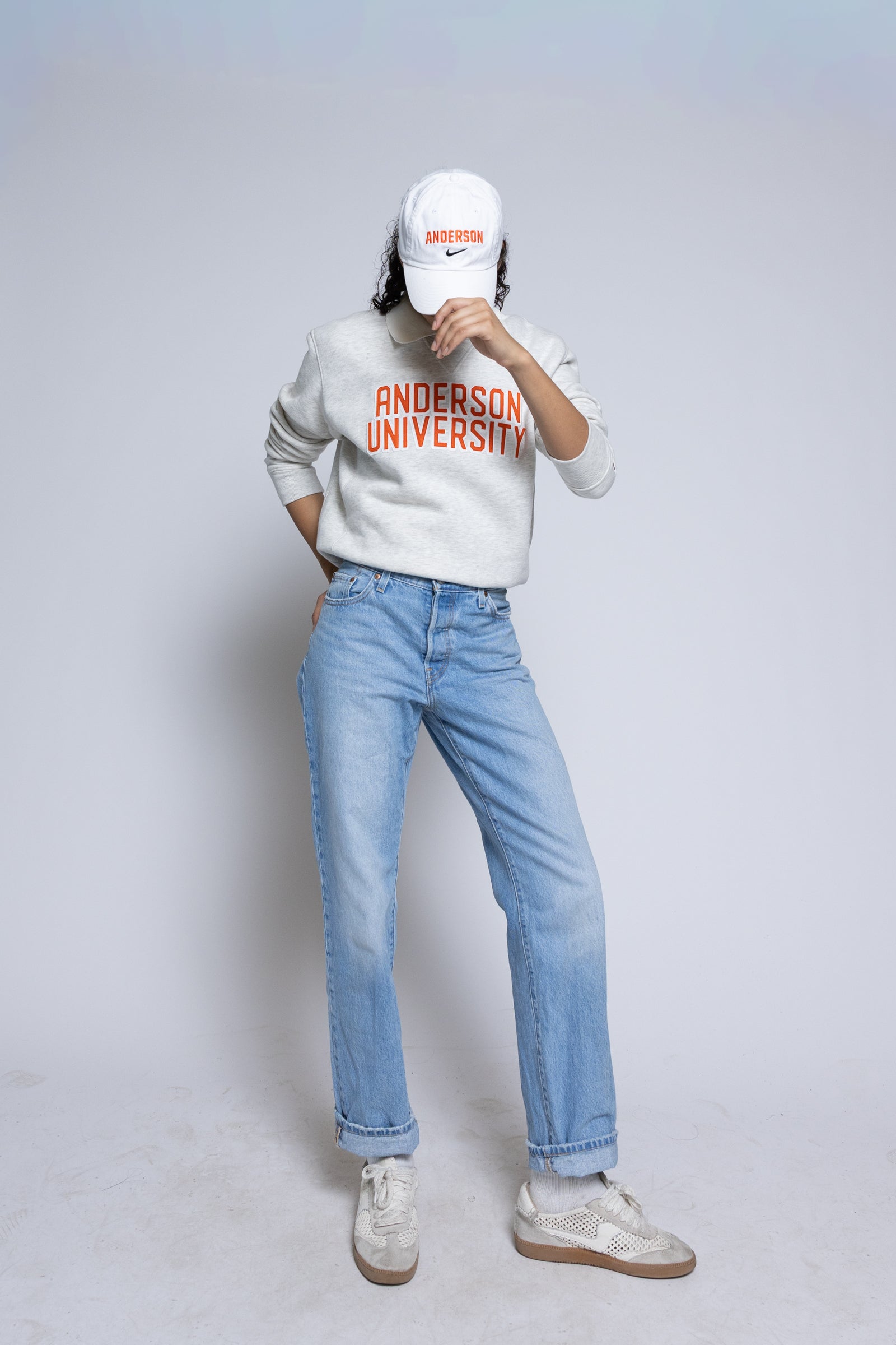 Person wearing an 'Anderson University' sweatshirt with a white cap and light blue jeans on a plain background